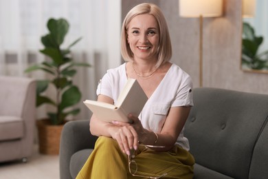 Portrait of smiling middle aged woman with glasses reading book on sofa at home