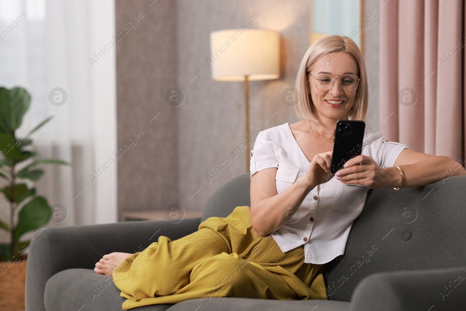 Photo of Portrait of smiling middle aged woman with smartphone on sofa at home. Space for text