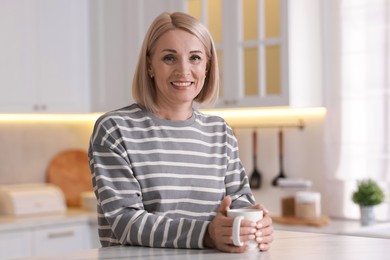 Smiling middle aged woman with cup of hot drink in kitchen