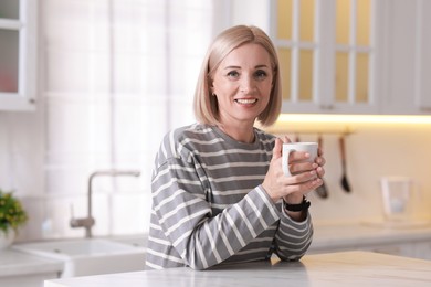 Smiling middle aged woman with cup of hot drink in kitchen