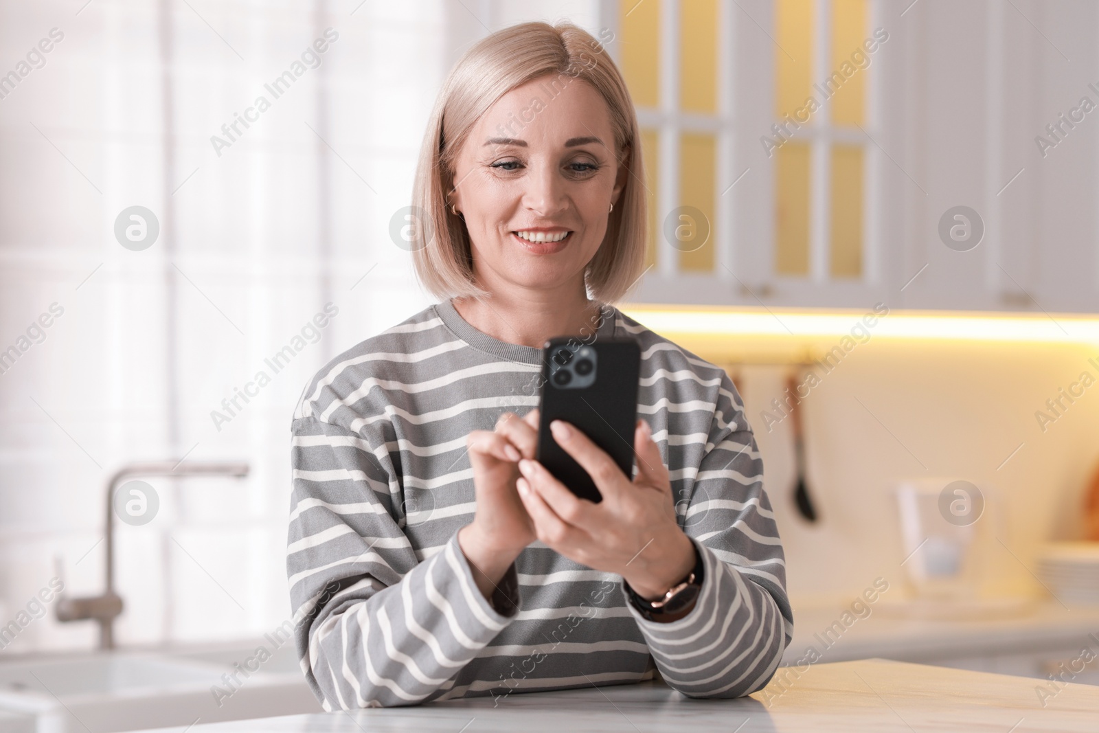 Photo of Portrait of smiling middle aged woman using smartphone in kitchen