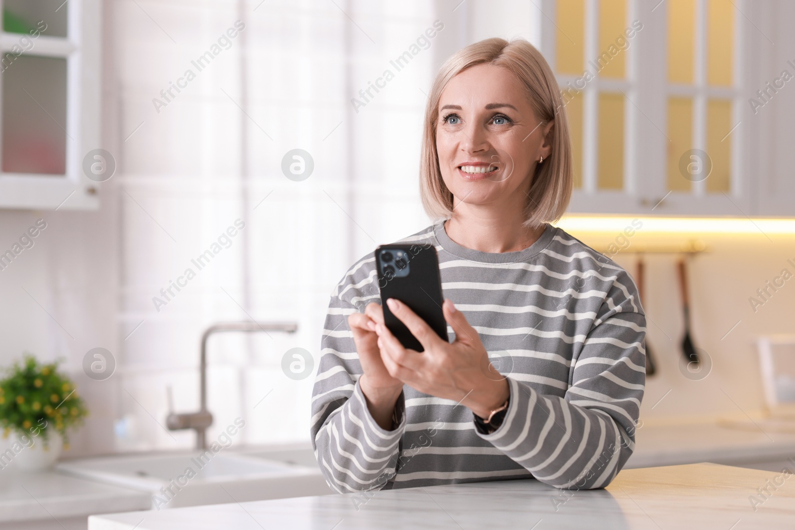 Photo of Portrait of smiling middle aged woman using smartphone in kitchen. Space for text