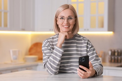 Photo of Portrait of smiling middle aged woman with smartphone in kitchen