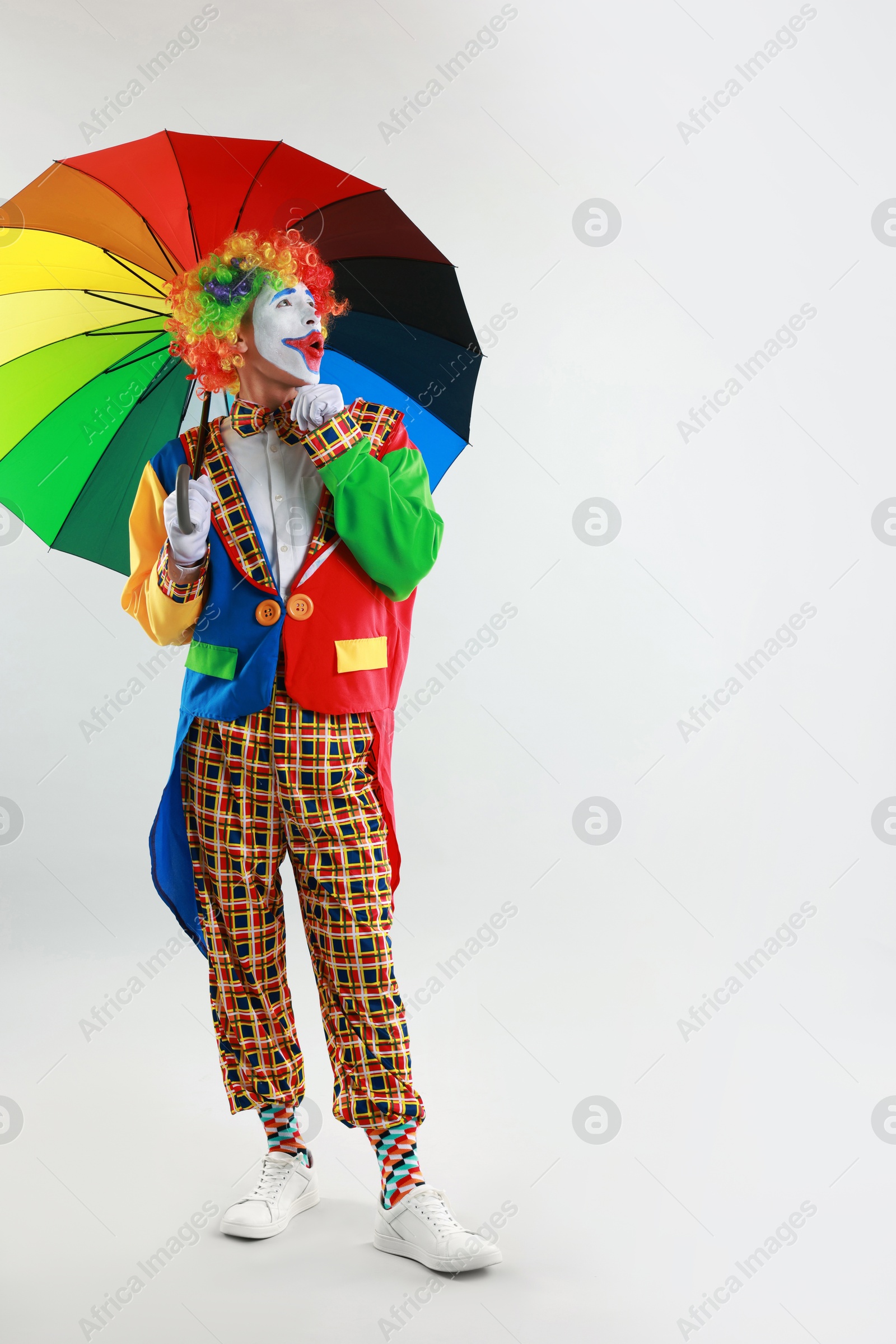 Photo of Emotional clown with colorful umbrella on light background, space for text