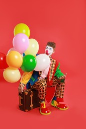 Clown with colorful balloons and suitcase on red background