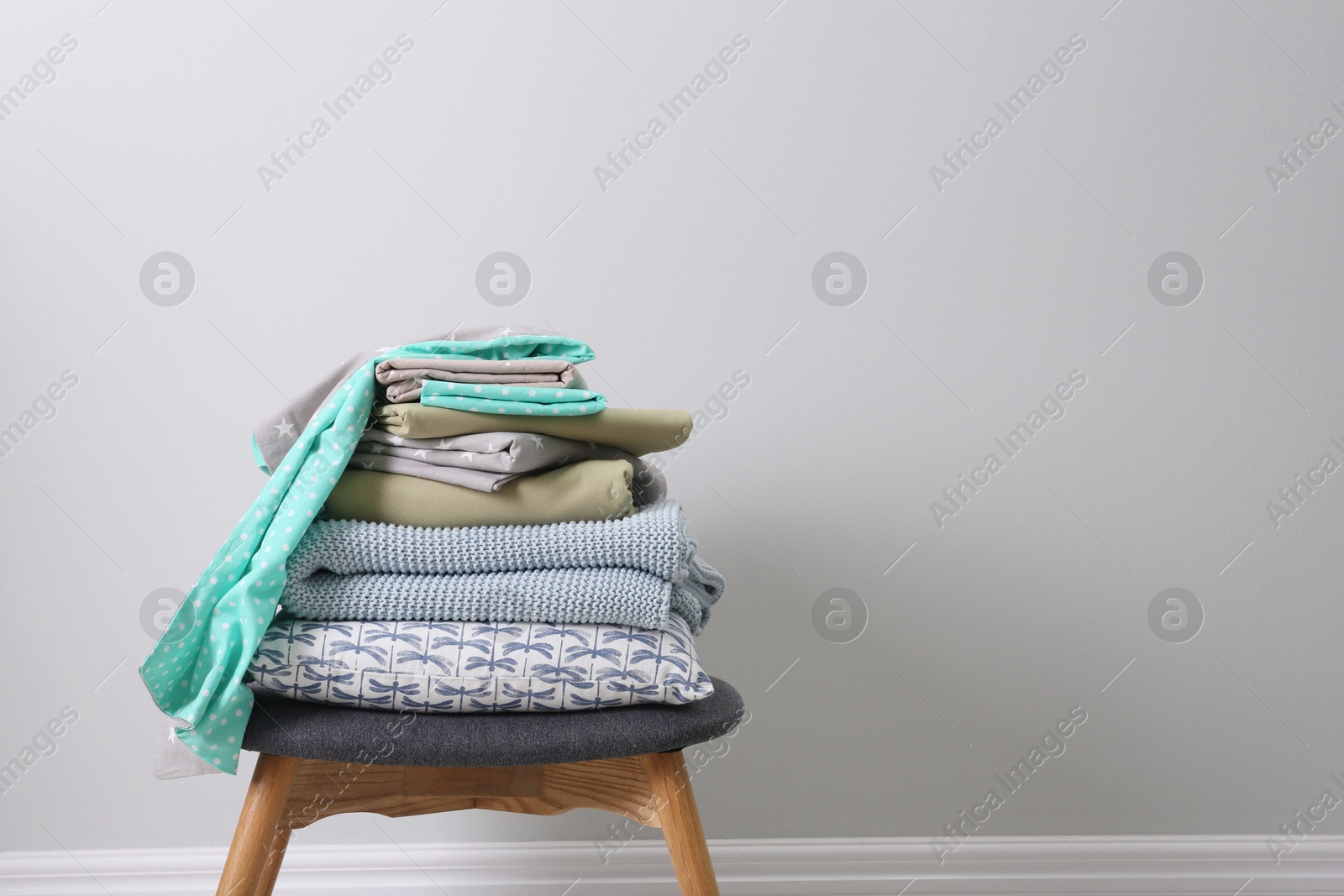 Photo of Stack of clean bed linen, plaid and pillow on stool near light grey wall. Space for text