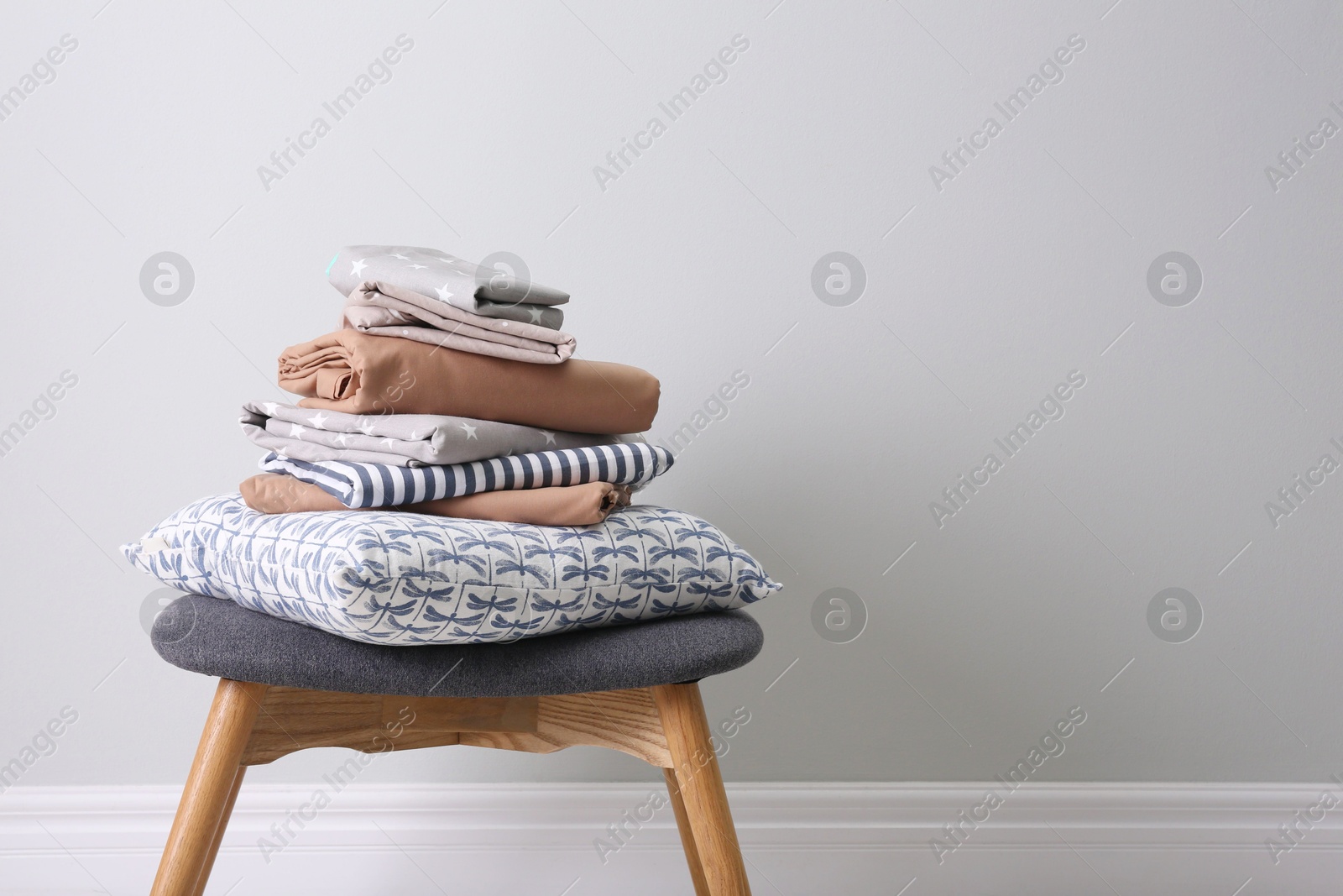 Photo of Stack of clean bed linen and pillow on stool near light grey wall. Space for text