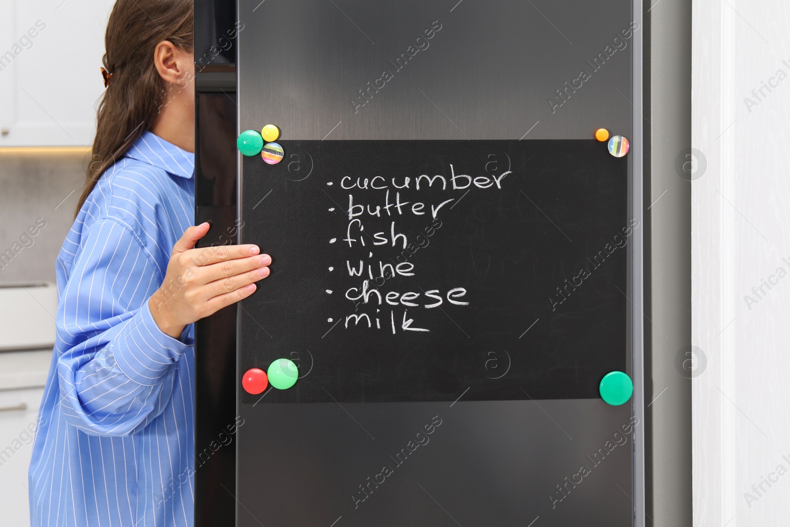 Photo of Woman opening refrigerator with shopping list on magnetic board in kitchen, closeup