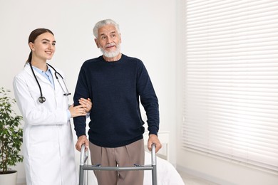 Doctor helping senior man with walking frame in clinic. Space for text