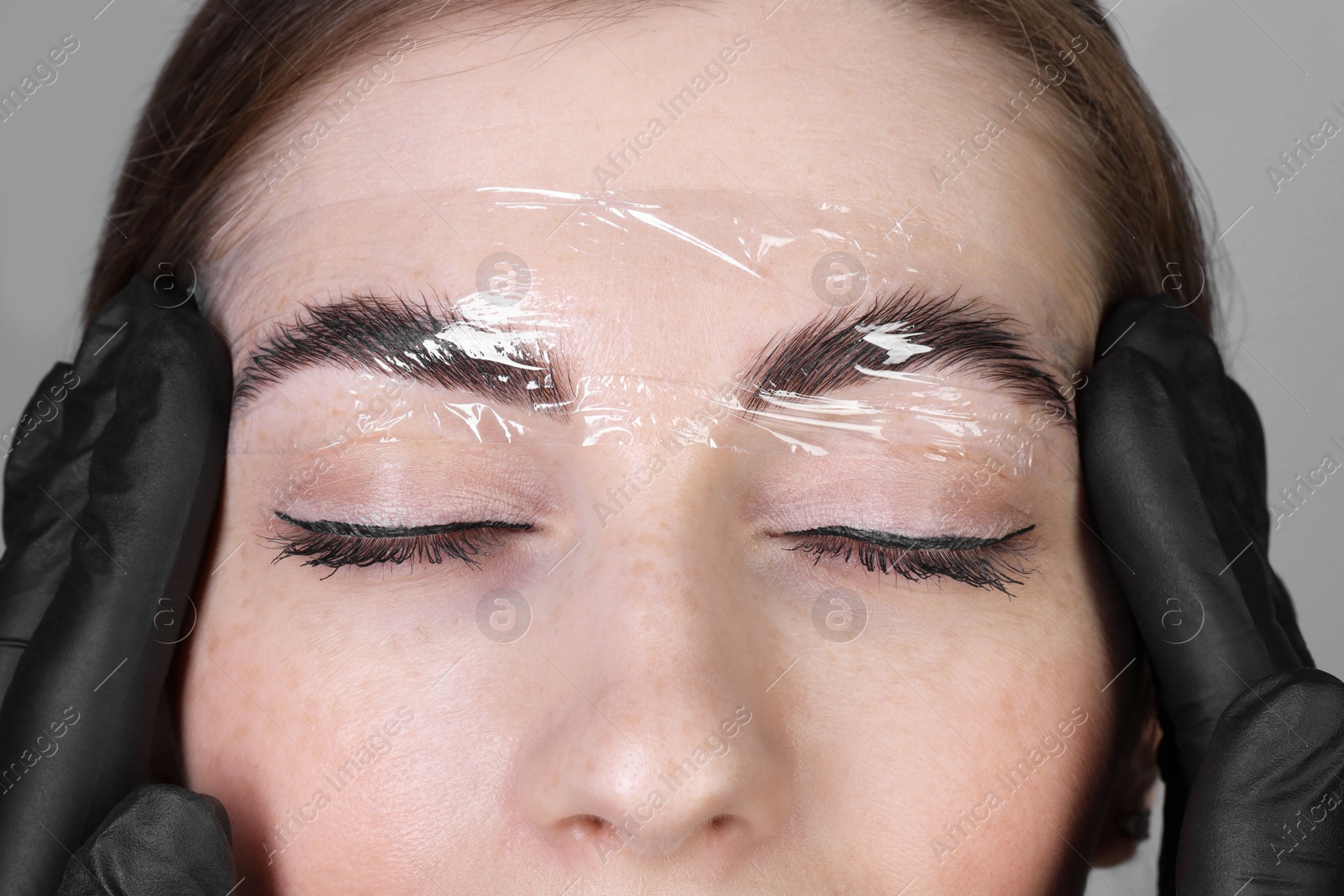 Photo of Brow lamination. Cosmetologist putting film onto woman's eyebrows against grey background, closeup