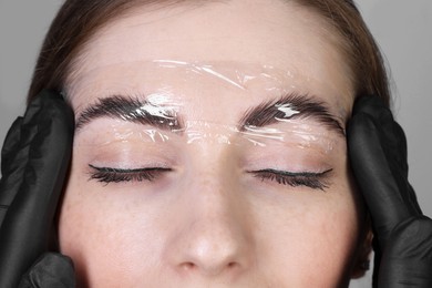 Brow lamination. Cosmetologist putting film onto woman's eyebrows against grey background, closeup