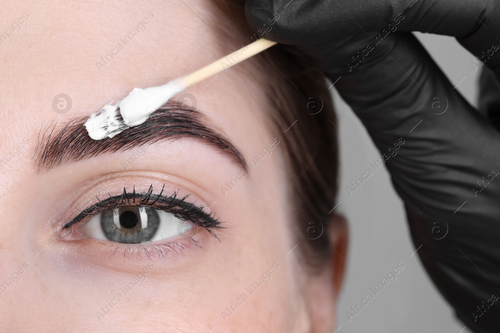 Photo of Brow lamination. Cosmetologist applying cream onto woman's eyebrows, closeup