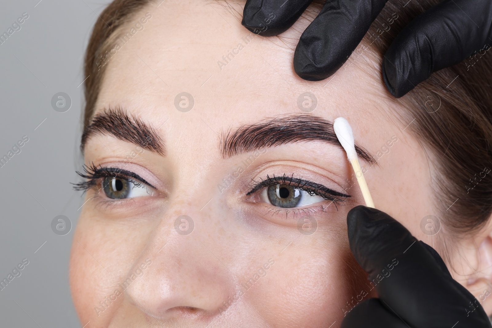 Photo of Brow lamination. Cosmetologist applying cream onto woman's eyebrows against grey background, closeup