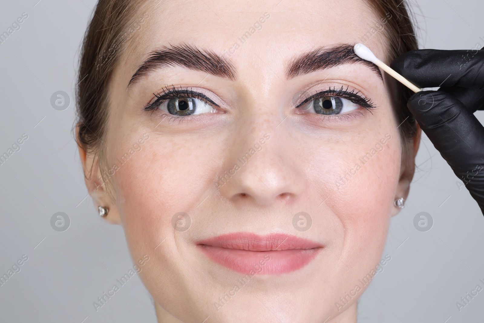 Photo of Brow lamination. Cosmetologist applying cream onto woman's eyebrows against grey background, closeup