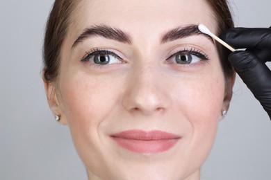 Photo of Brow lamination. Cosmetologist applying cream onto woman's eyebrows against grey background, closeup