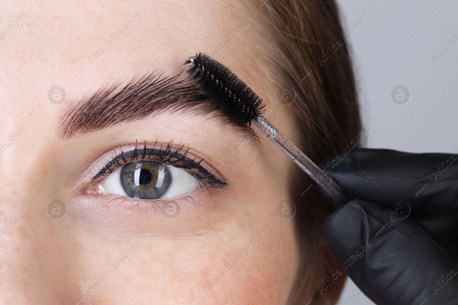 Photo of Brow lamination. Cosmetologist combing woman's eyebrows with brush against grey background, closeup