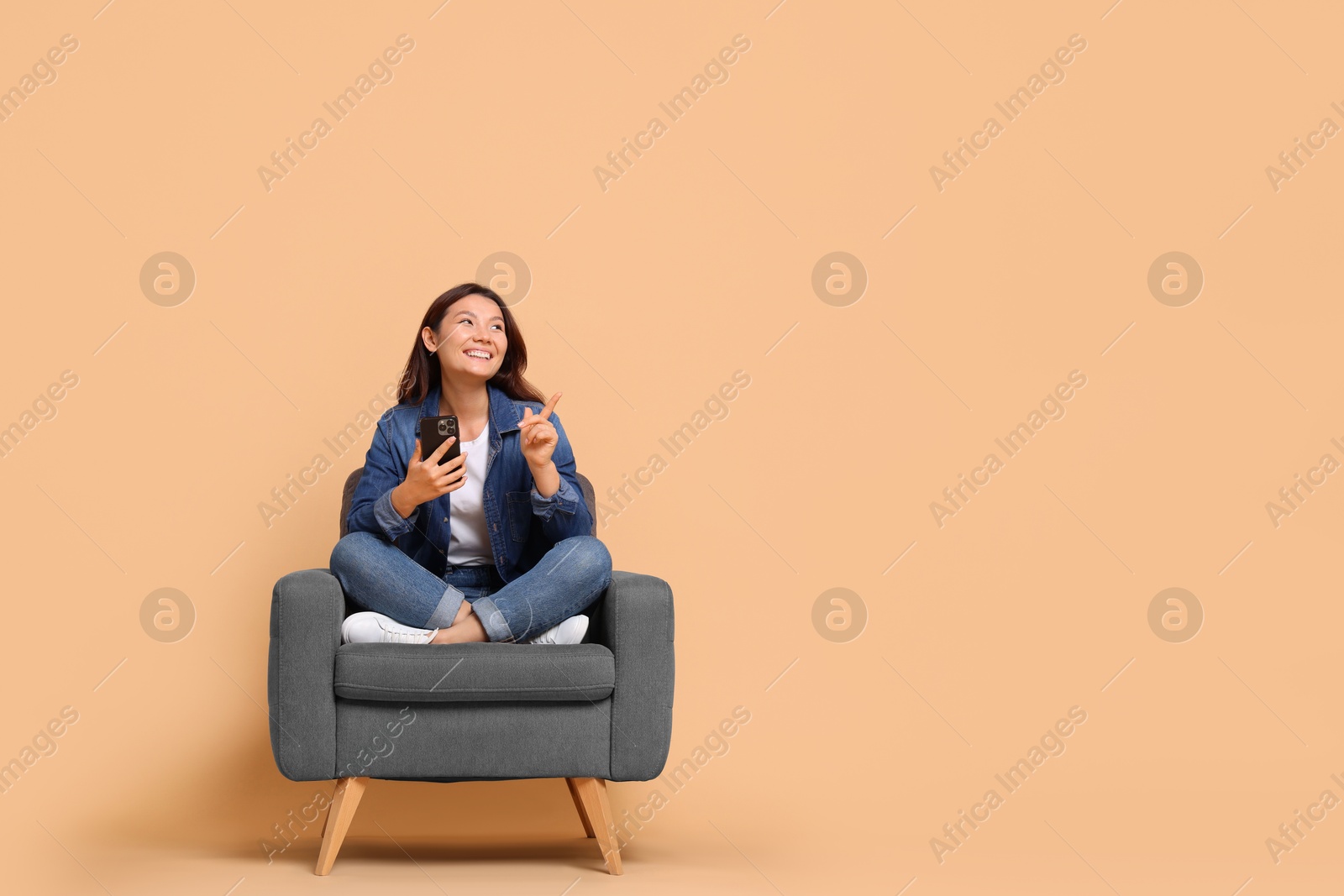 Photo of Smiling woman with smartphone sitting in armchair on beige background. Space for text