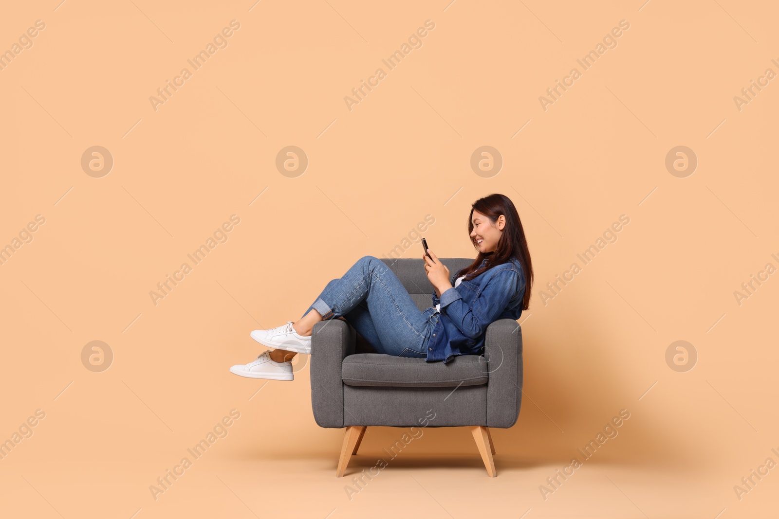 Photo of Smiling woman with smartphone sitting in armchair on beige background