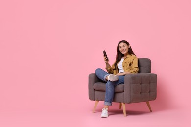 Smiling woman with smartphone sitting in armchair on pink background. Space for text