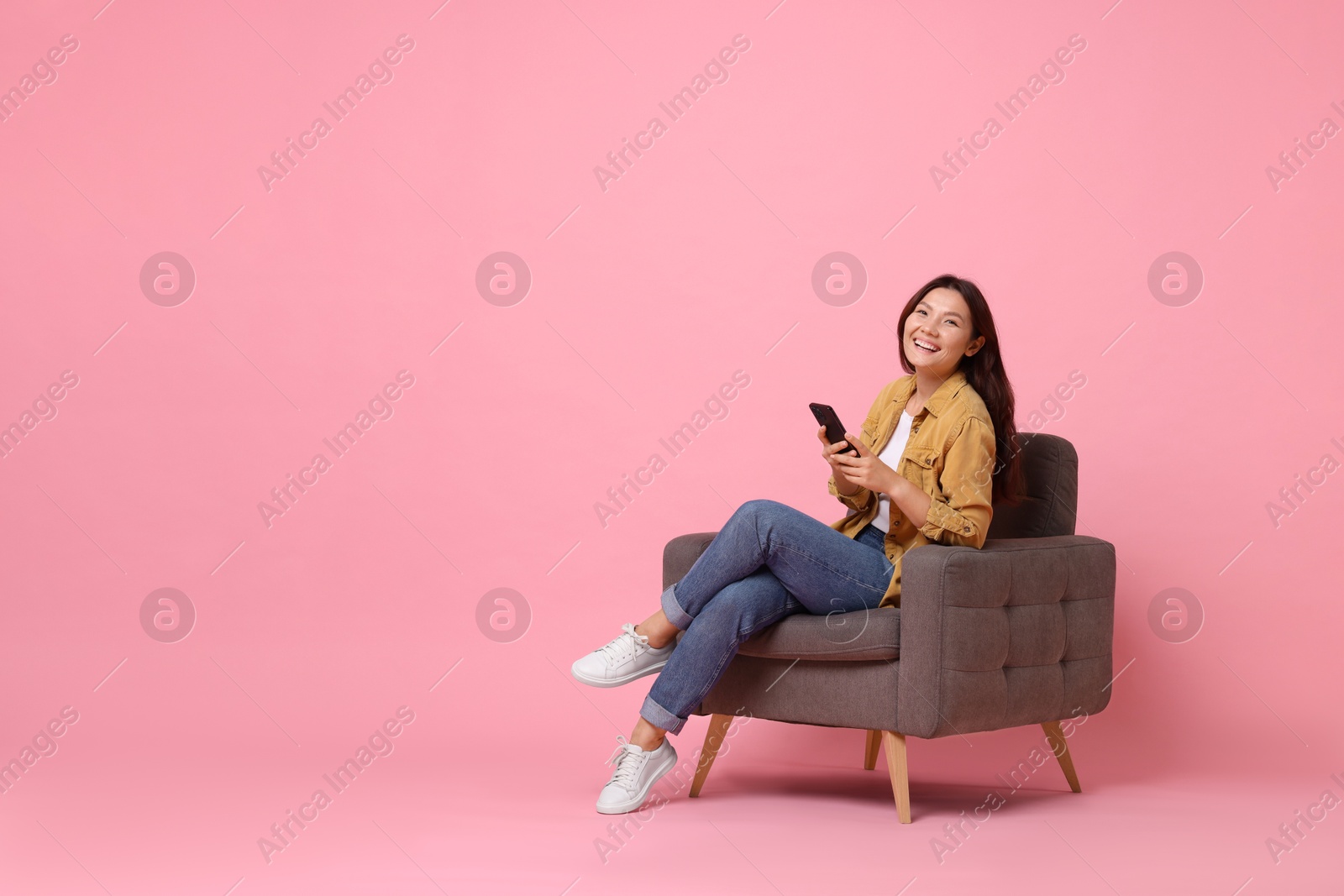 Photo of Smiling woman with smartphone sitting in armchair on pink background. Space for text