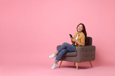 Smiling woman with smartphone sitting in armchair on pink background. Space for text