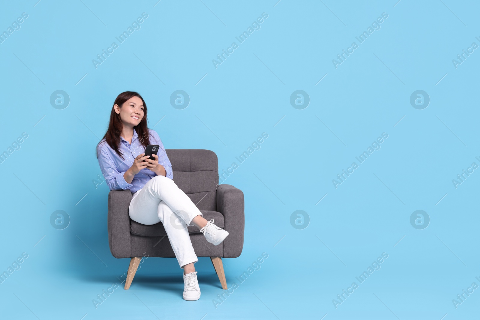 Photo of Smiling woman with smartphone resting in armchair on light blue background. Space for text