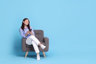 Smiling woman with smartphone resting in armchair on light blue background. Space for text