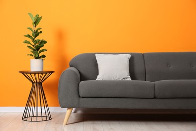 Photo of Stylish sofa, pillow and side table with houseplant near orange wall indoors