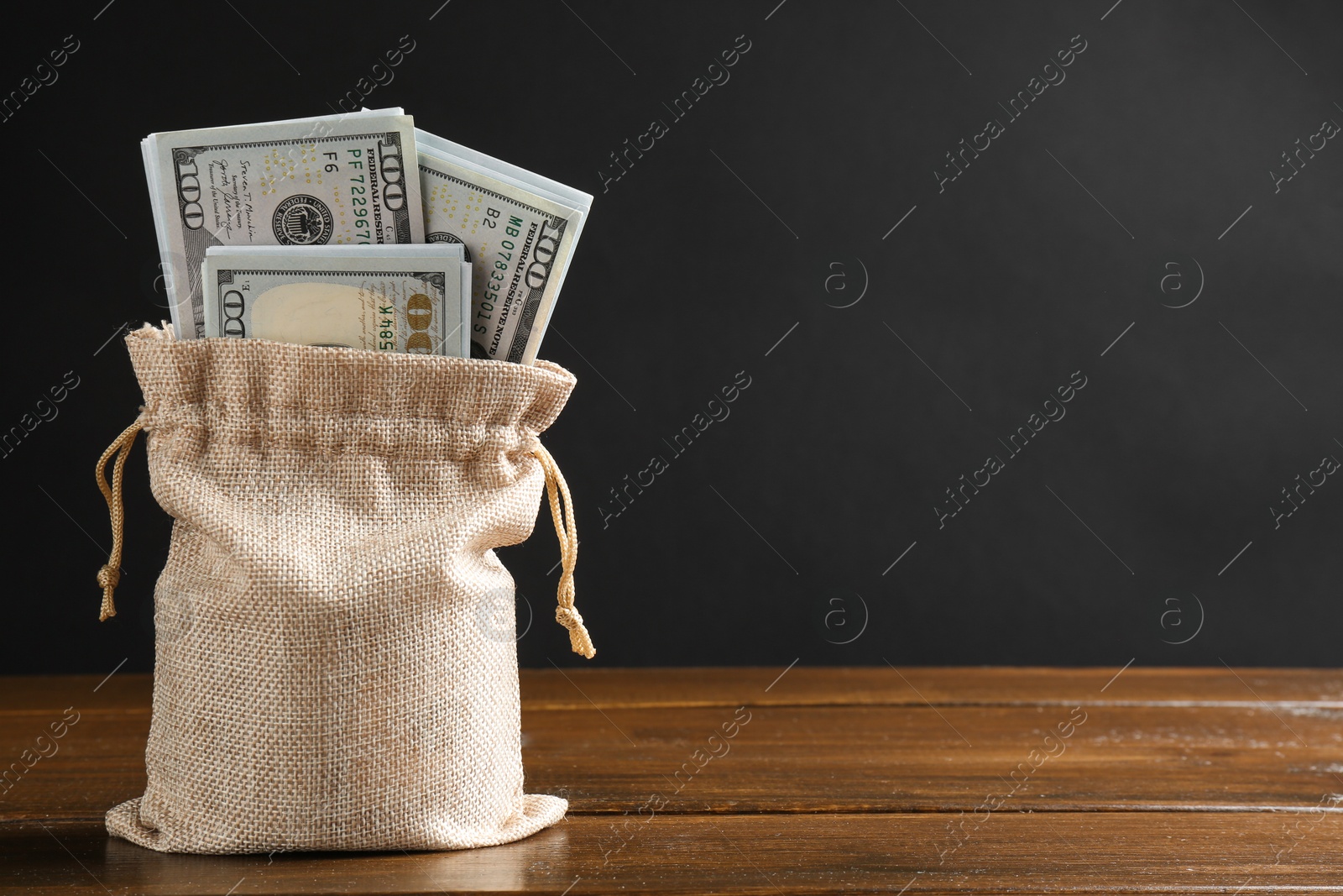 Photo of Dollar banknotes in burlap sack on wooden table against dark background, space for text