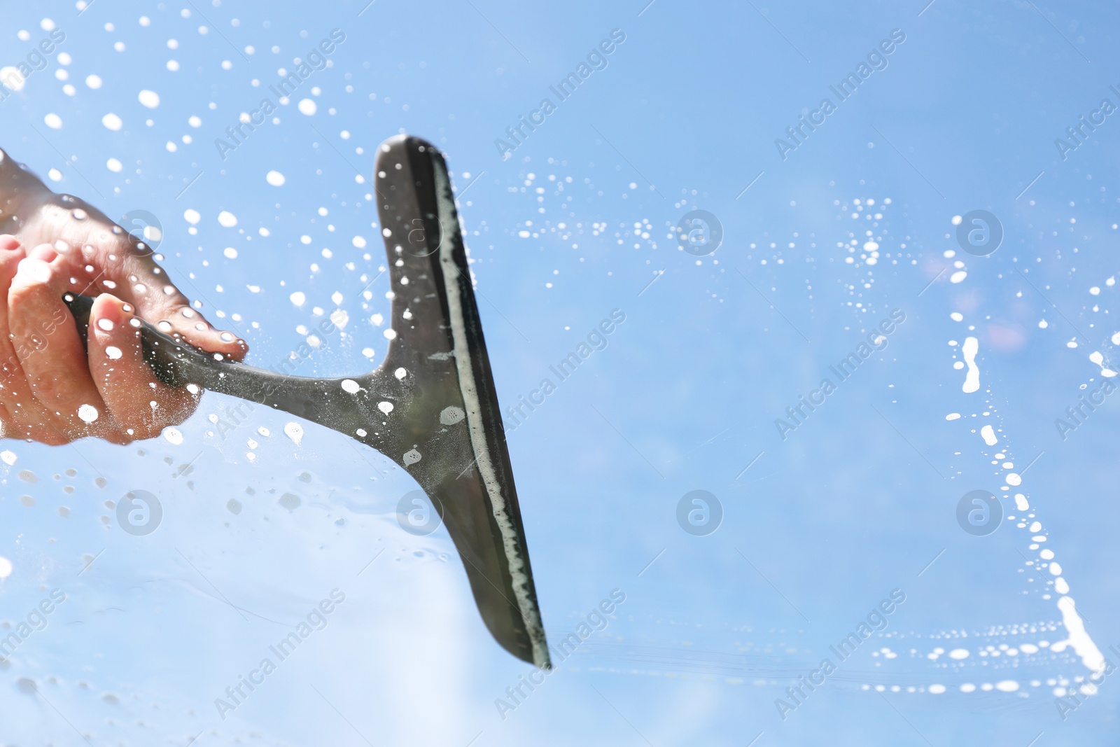 Photo of Woman washing window with squeegee tool against blue sky, closeup