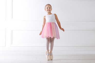 Photo of Little ballerina practicing dance moves near white wall indoors