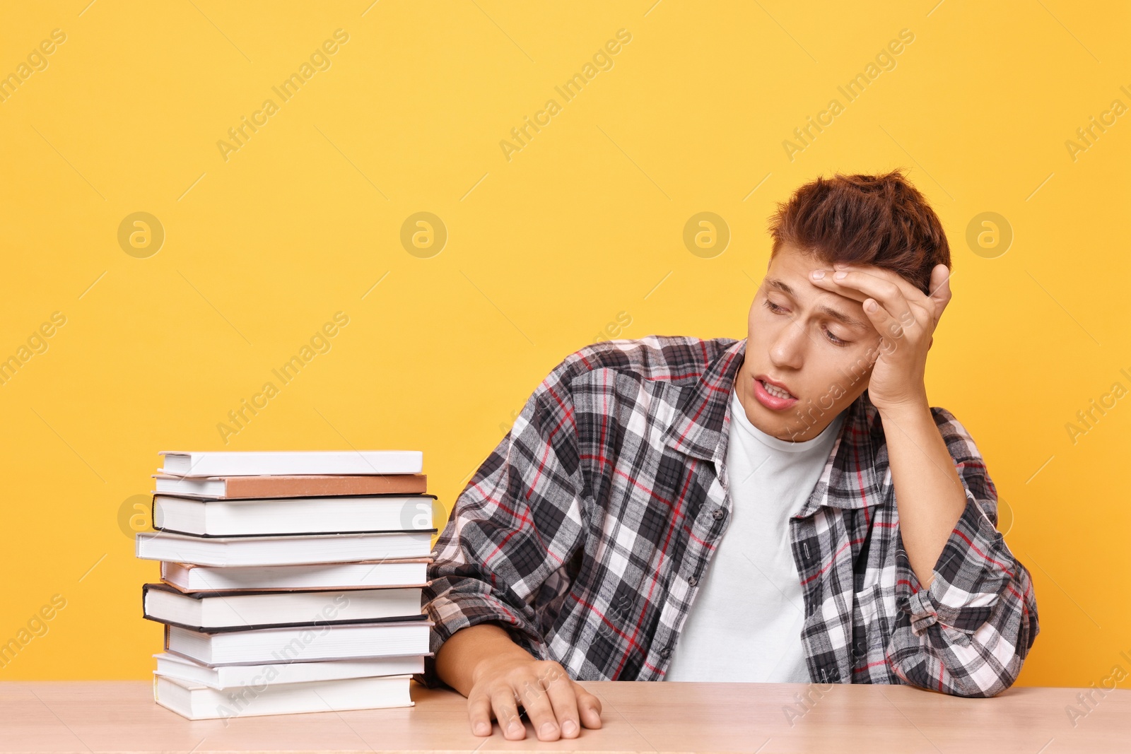 Photo of Young student with stack of books having stress before exam at wooden table against yellow background