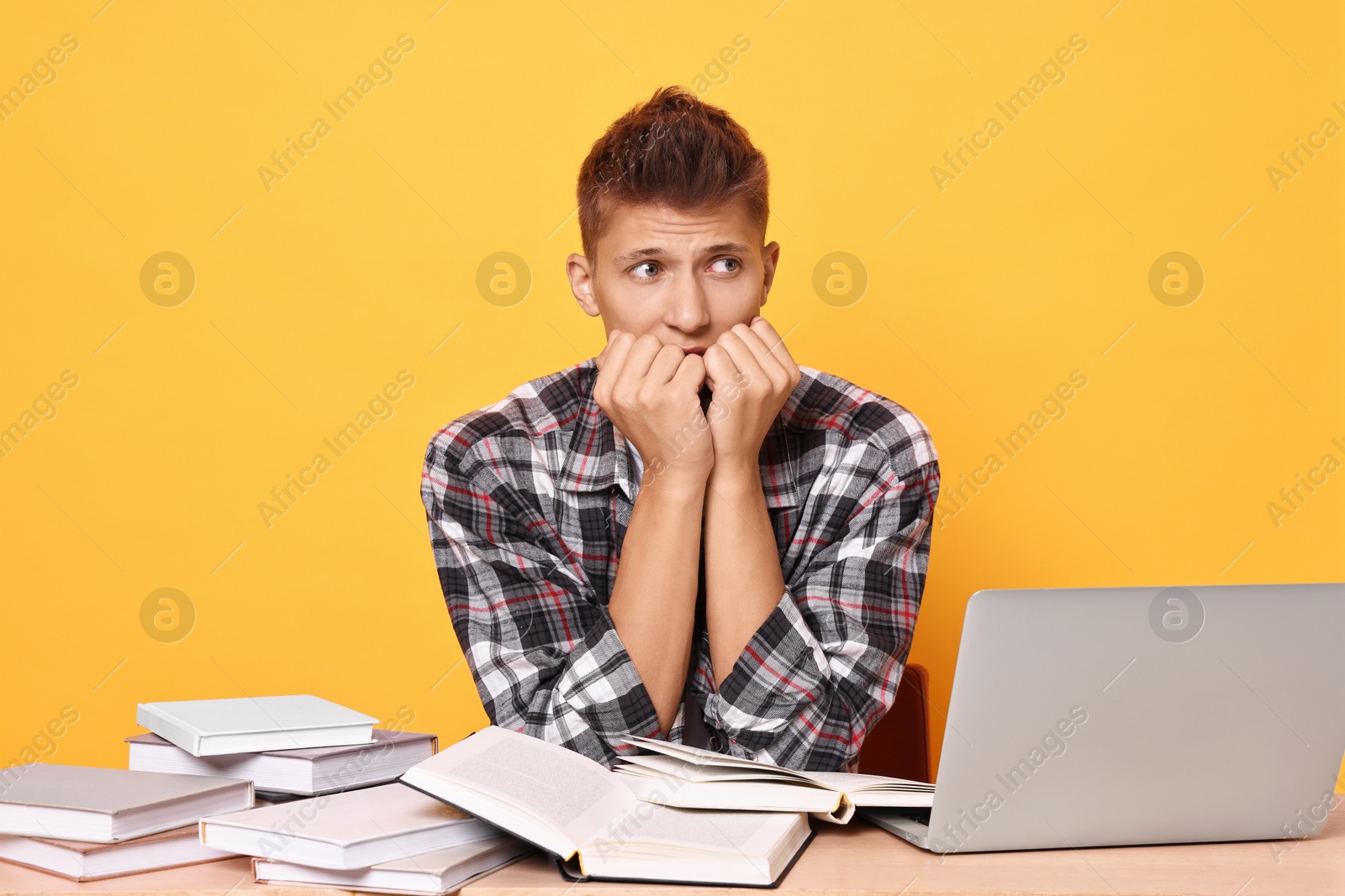 Photo of Young student with books and laptop having stress before exam at table against yellow background