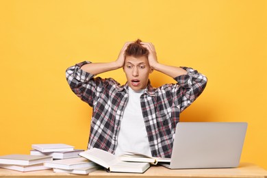 Photo of Emotional student with books and laptop having stress before exam at table against yellow background