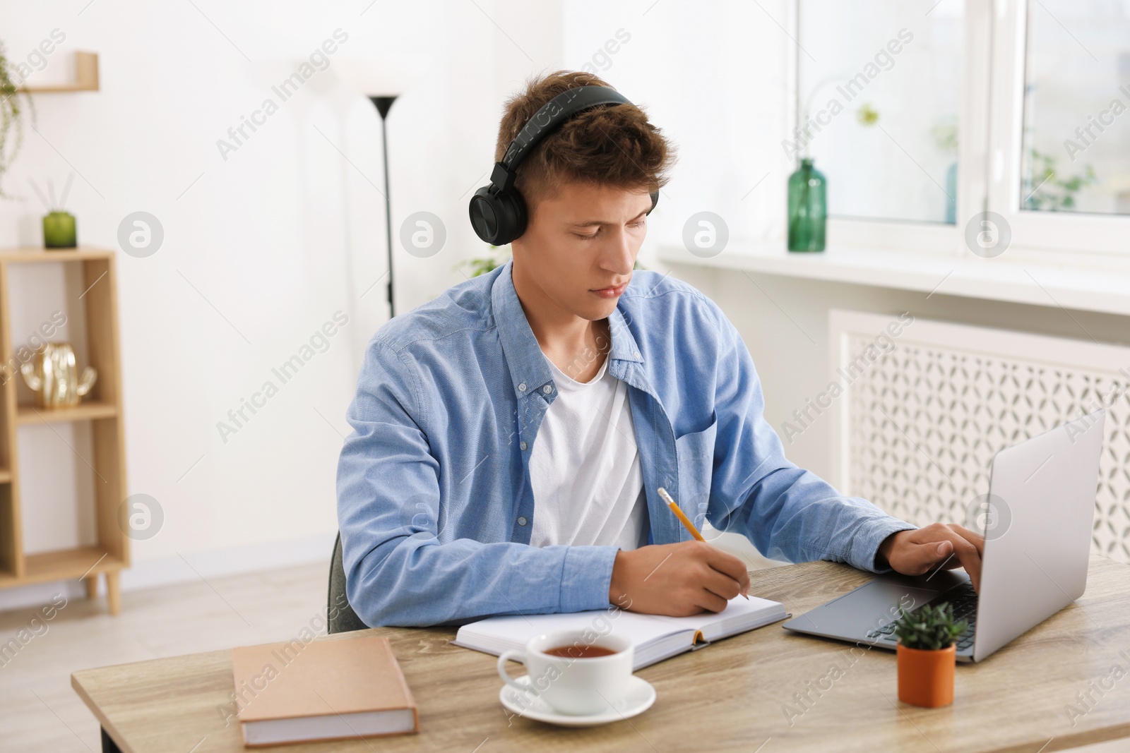 Photo of Student in headphones taking notes while studying with laptop at wooden table indoors