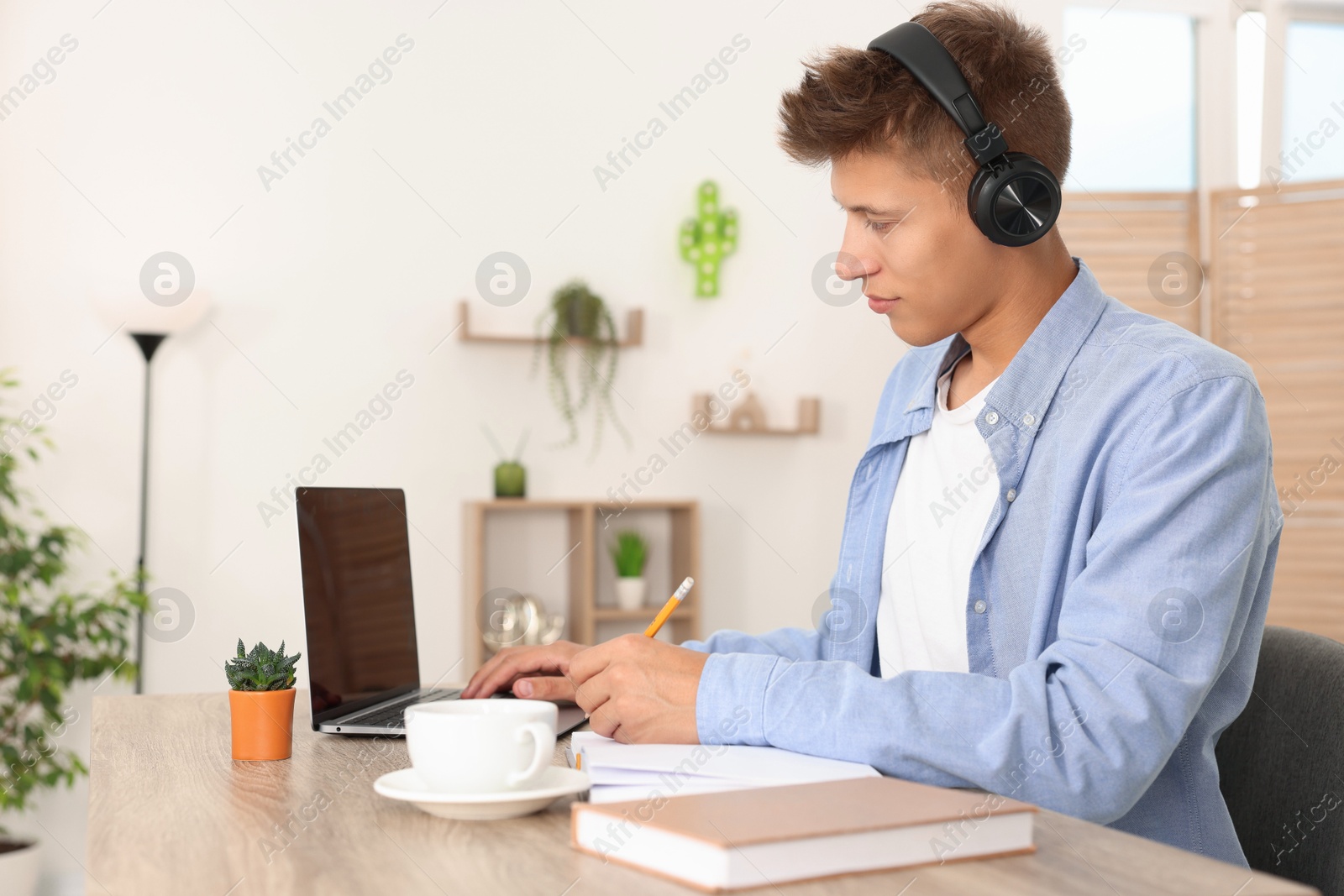 Photo of Student in headphones taking notes while studying with laptop at wooden table indoors