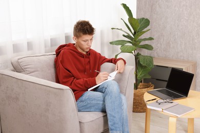 Student taking notes while studying in armchair at home