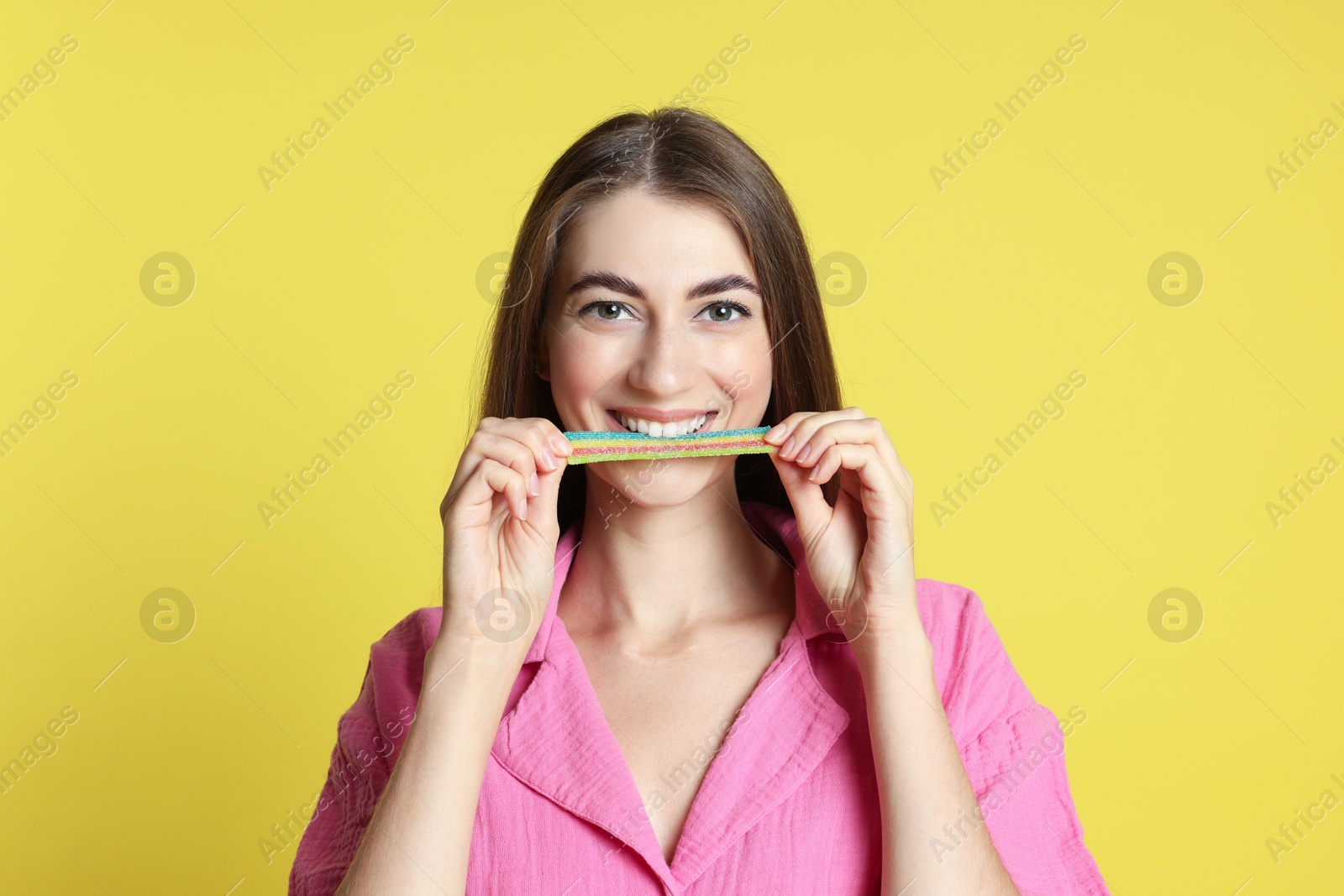 Photo of Young woman eating tasty rainbow sour belt on yellow background