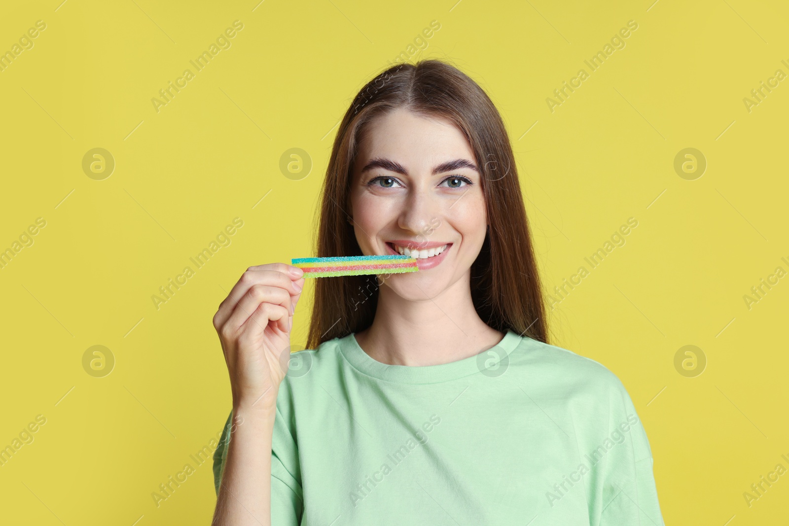 Photo of Young woman eating tasty rainbow sour belt on yellow background