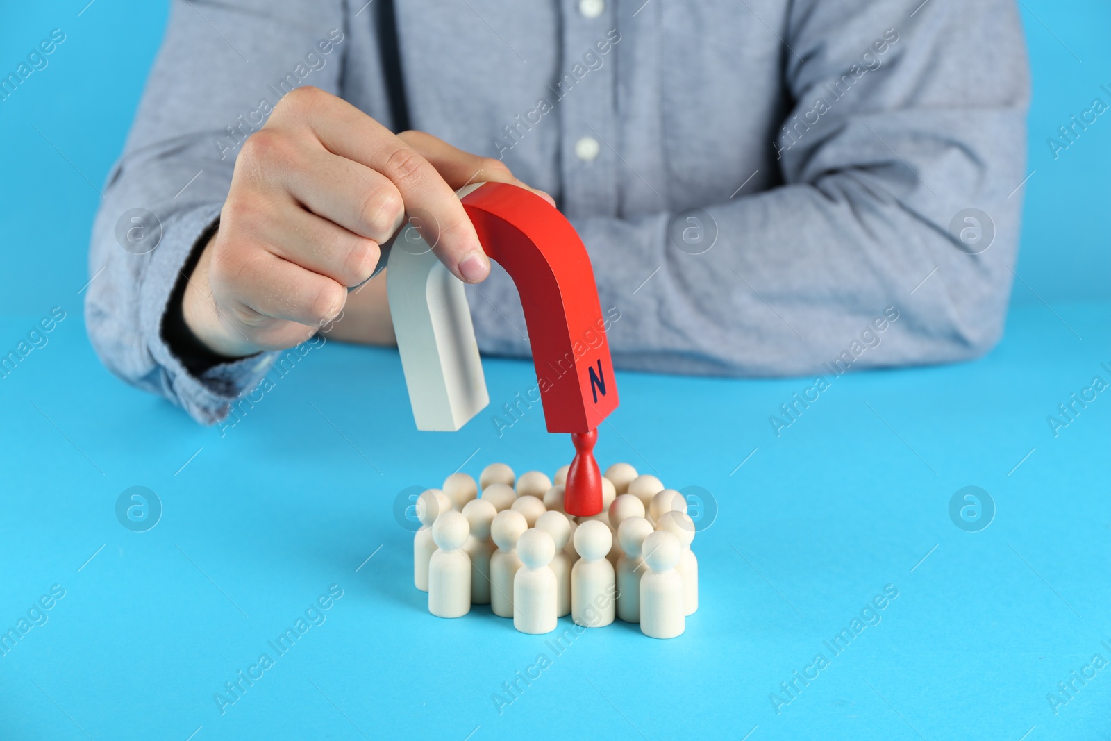 Photo of Man with magnet attracting red piece among wooden ones on light blue background, closeup