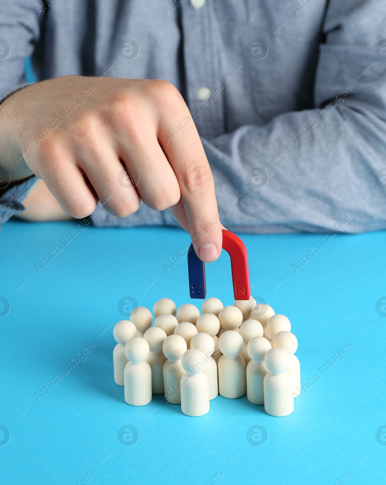 Photo of Man with magnet attracting wooden human figures on light blue background, closeup