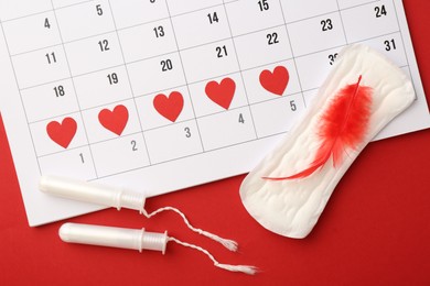 Photo of Calendar with marked dates, menstrual pad with feather and tampons on red background, top view