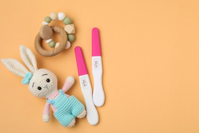 Photo of Pregnancy tests, baby rattle and toy bunny on pale orange background, top view. Space for text