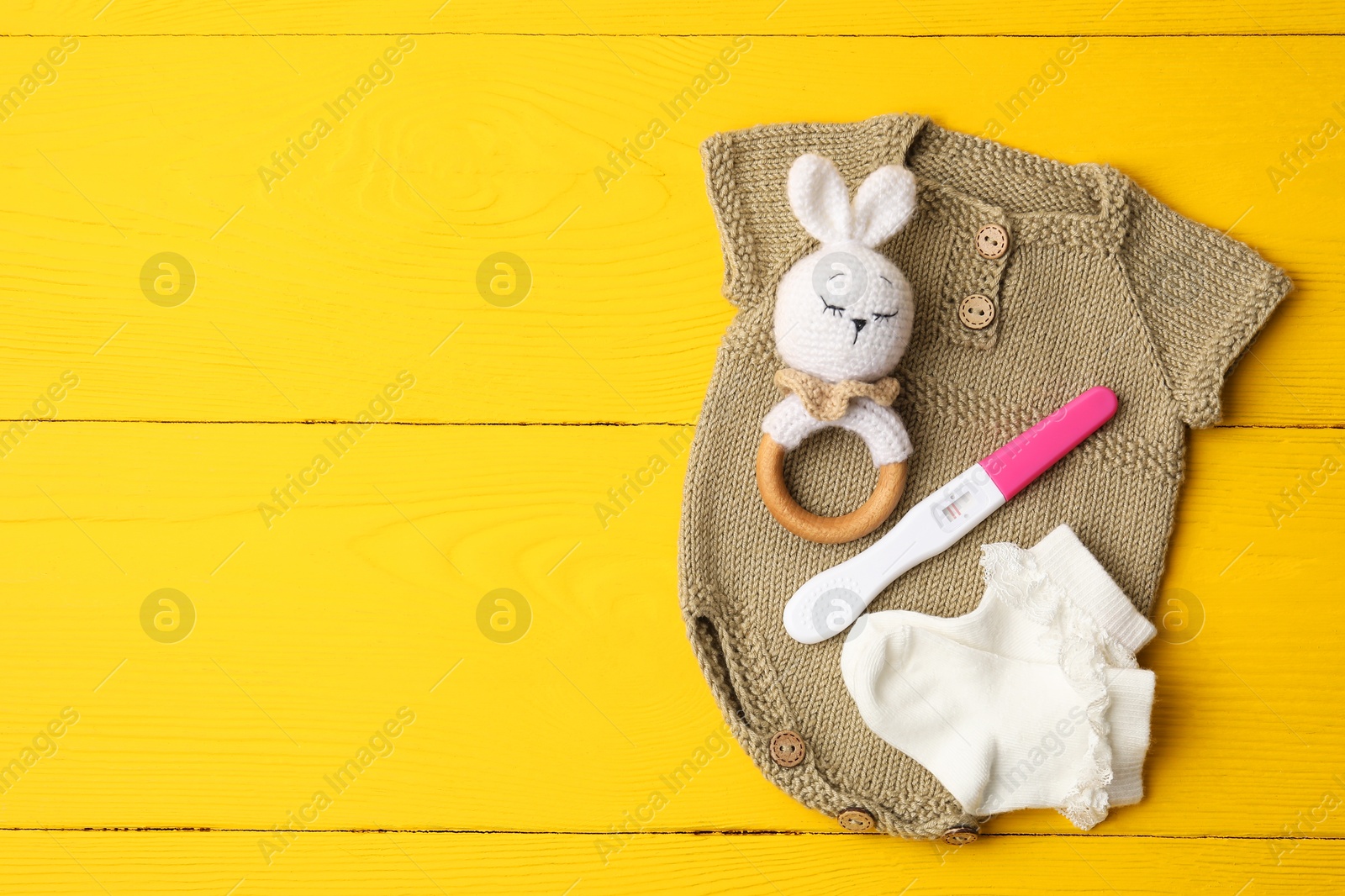 Photo of Pregnancy test, baby onesie, toy and socks on yellow wooden table, flat lay. Space for text