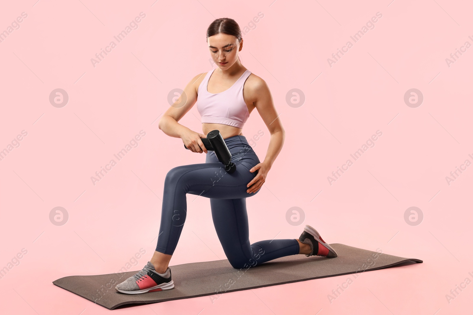 Photo of Woman using percussive massager to relax hip muscles on mat against pink background