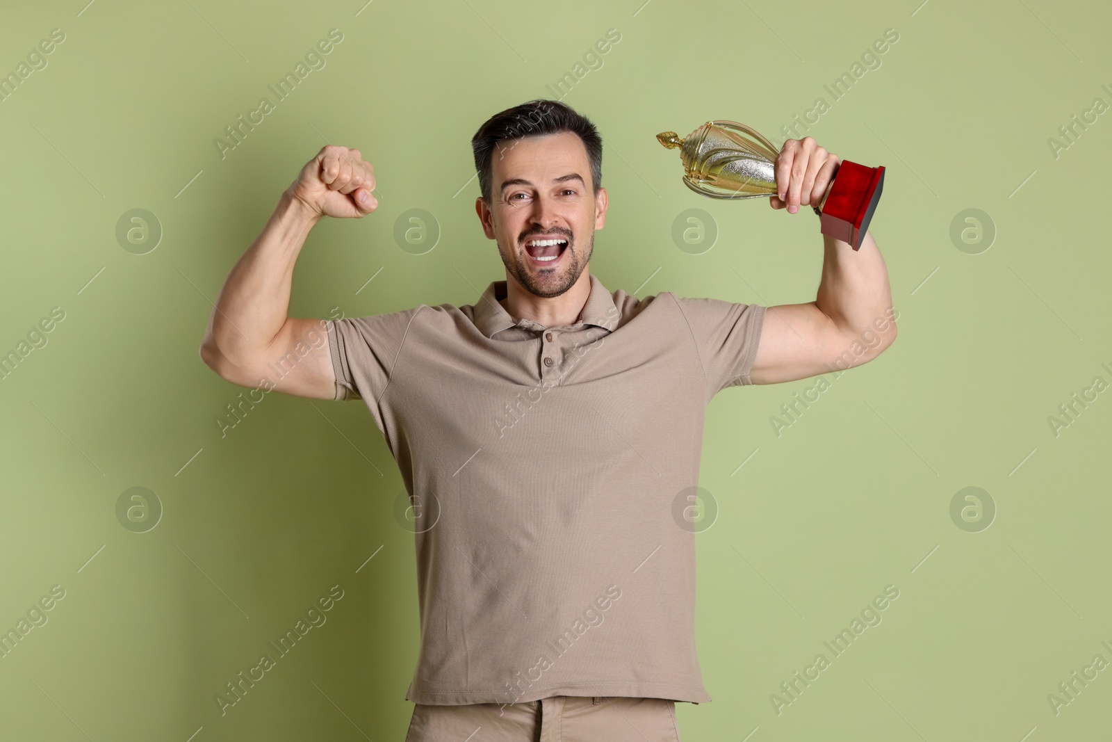 Photo of Happy winner with golden trophy cup showing his biceps on pale olive background