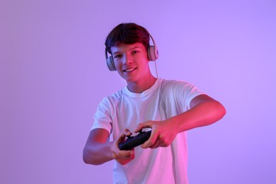 Smiling teenage boy in headphones playing video game with controller on violet background