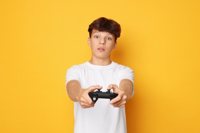 Emotional teenage boy playing video game with controller on orange background