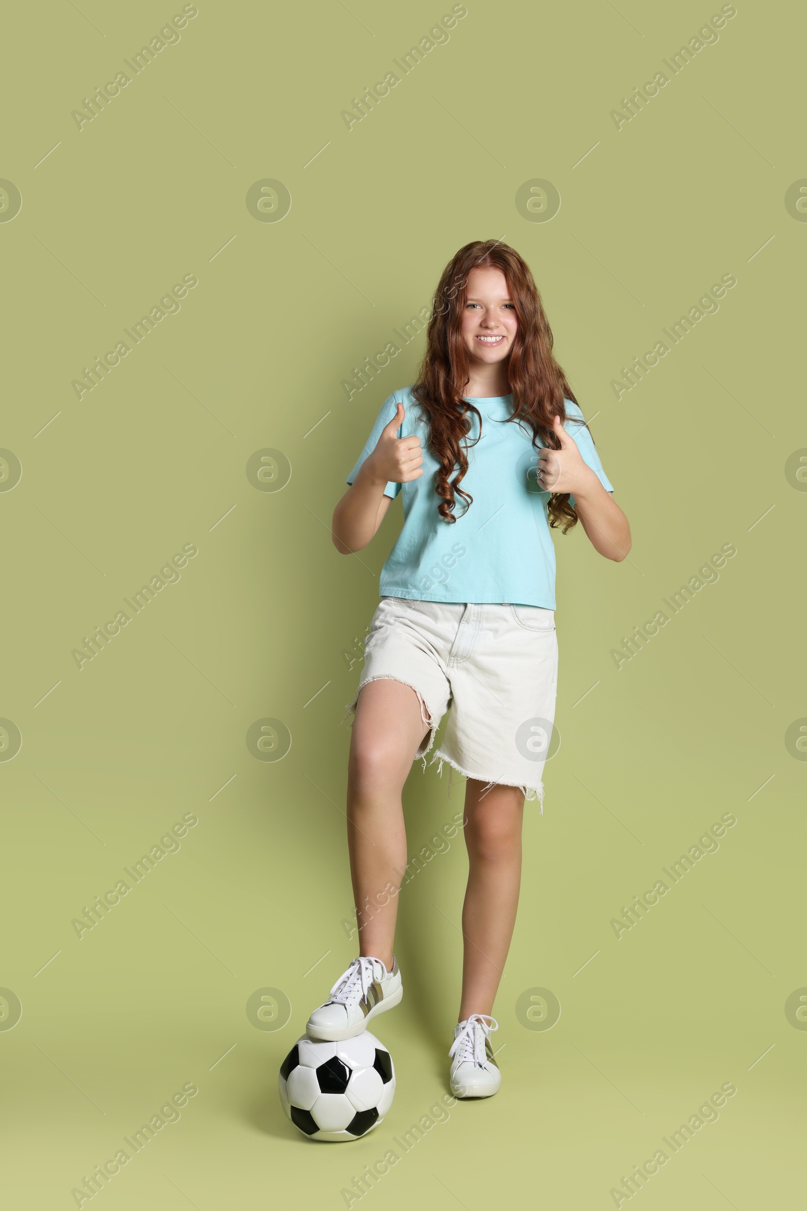 Photo of Smiling teenage girl with soccer ball showing thumbs up on green background