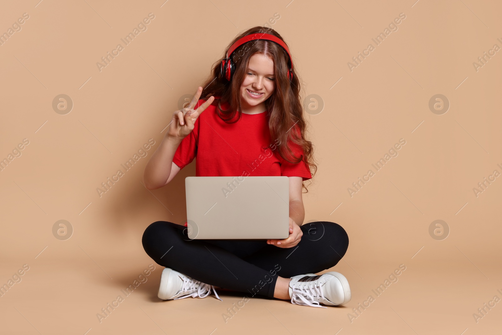 Photo of Smiling teenage girl in headphones showing peace sign with laptop on beige background